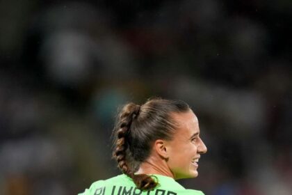 Female soccer player seen from behind in a bright green kit with 'PLUMPTRE' and number 2, smiling on the field