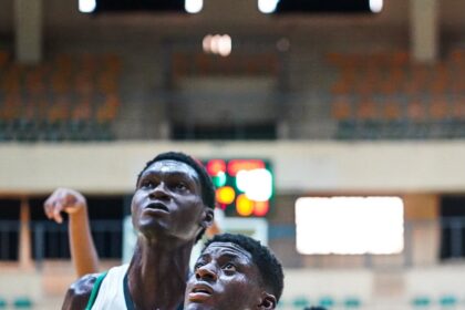 Two basketball players under a hoop fight for position, Nigeria jersey number 8 in green foreground, defender in white behind, indoors arena with blurred scoreboard in background