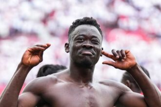 Muscular shirtless man posing with arms raised at a stadium, crowd blurred in the background, celebrating.