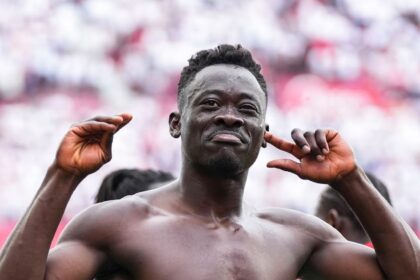 Muscular shirtless man posing with arms raised at a stadium, crowd blurred in the background, celebrating.