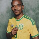 Portrait of a man in a yellow green soccer jersey with crest posed against a green textured background gripping the shirt at chest level  trybalnews