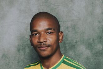 Portrait of a man in a yellow-green soccer jersey with crest, posed against a green textured background, gripping the shirt at chest level.