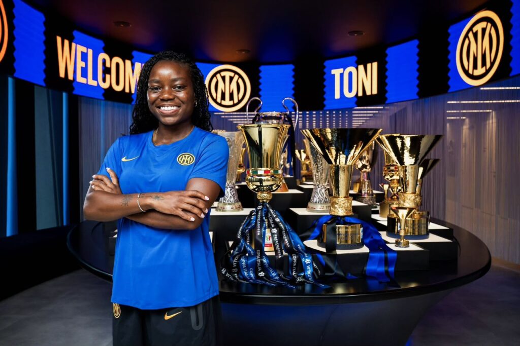Toni Payne Trybal News Smiling female athlete in a blue kit stands with arms crossed beside a display of shiny trophies and ribbons on a black pedestal trybalnews