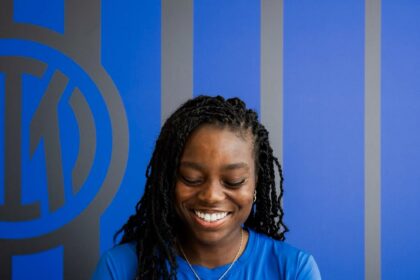 Smiling woman in a blue shirt signs documents at a white desk, wearing an Inter Milan badge on her chest.