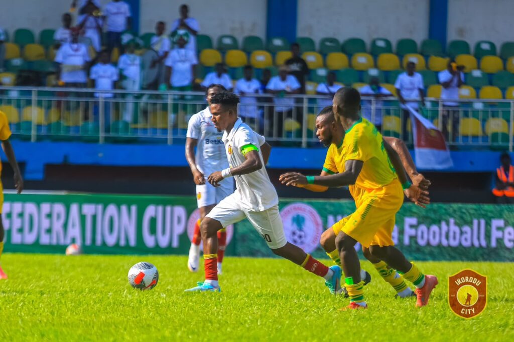 El Khanemi Vs Ikorodu City Trybal News Soccer player in white jersey with green armband dribbles the ball while two yellow clad opponents chase him on a sunny field stadium in background trybalnews