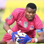 Goalkeeper in a pink kit and blue gloves catches a soccer ball during a match on a grassy field  trybalnews
