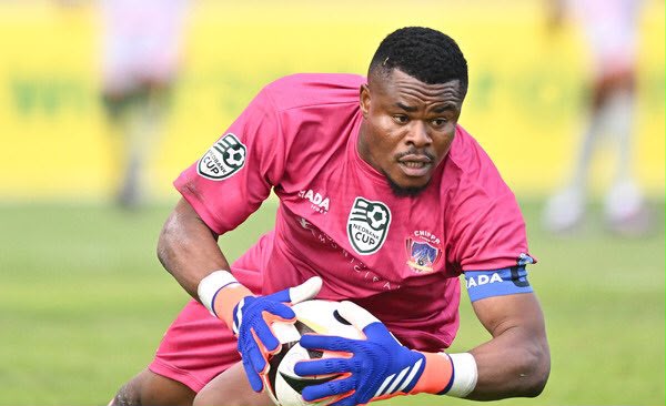Goalkeeper in a pink kit and blue gloves catches a soccer ball during a match on a grassy field  trybalnews