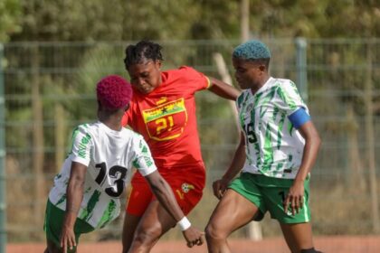Three soccer players compete for the ball during a match; a player in red is between two in white-and-green kits.