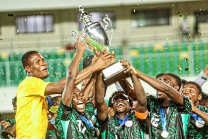 Soccer team in green and black celebrate holding a large silver trophy aloft, confetti flying.