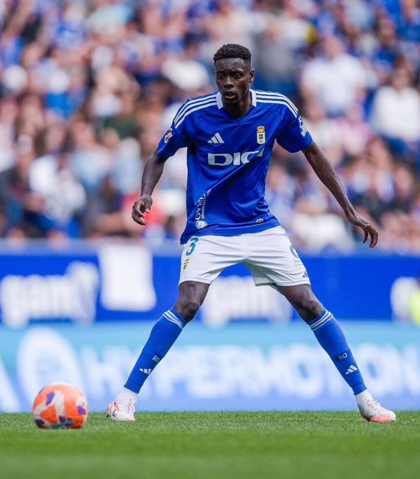 Footballer in a blue jersey and white shorts dribbles an orange ball on a green pitch with a blurred crowd in the background  trybalnews