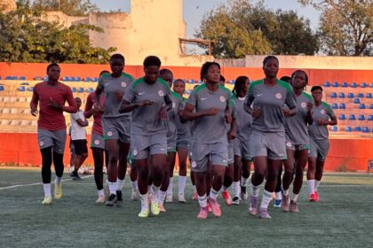 Group of female soccer players in gray uniforms jog on a field during training, with orange bleachers in the background.