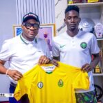 Two men in white polo shirts hold a bright yellow football jersey with logos in a trophy filled room  trybalnews