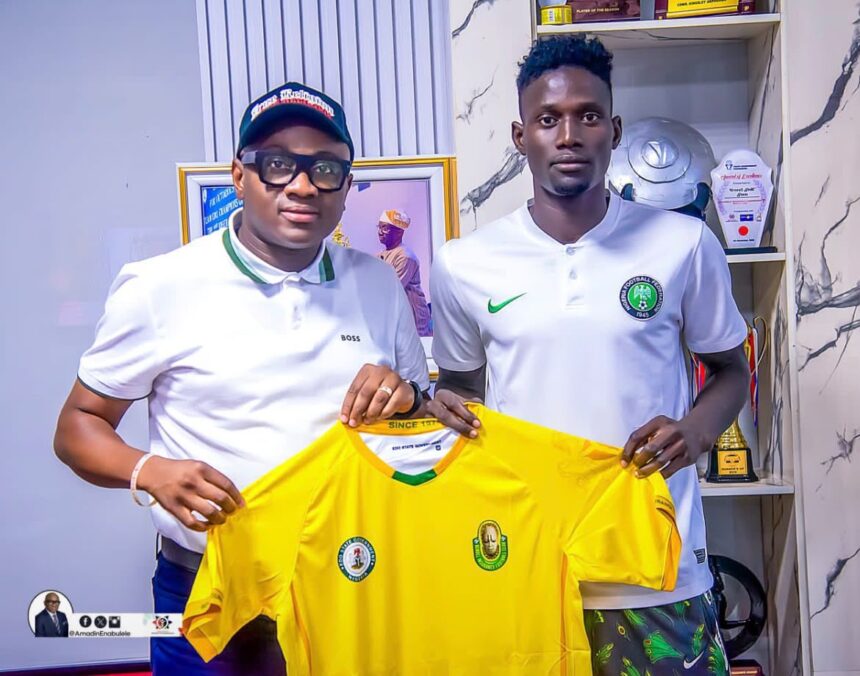 Two men in white polo shirts hold a bright yellow football jersey with logos in a trophy filled room  trybalnews