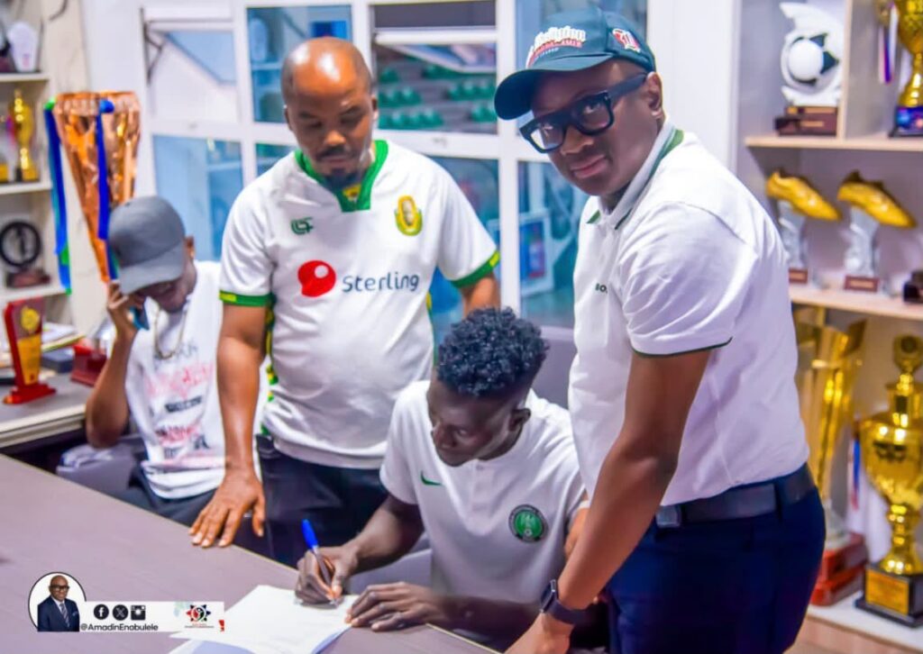 Group of young men in team jerseys sign a document at a table in a trophy filled room  trybalnews