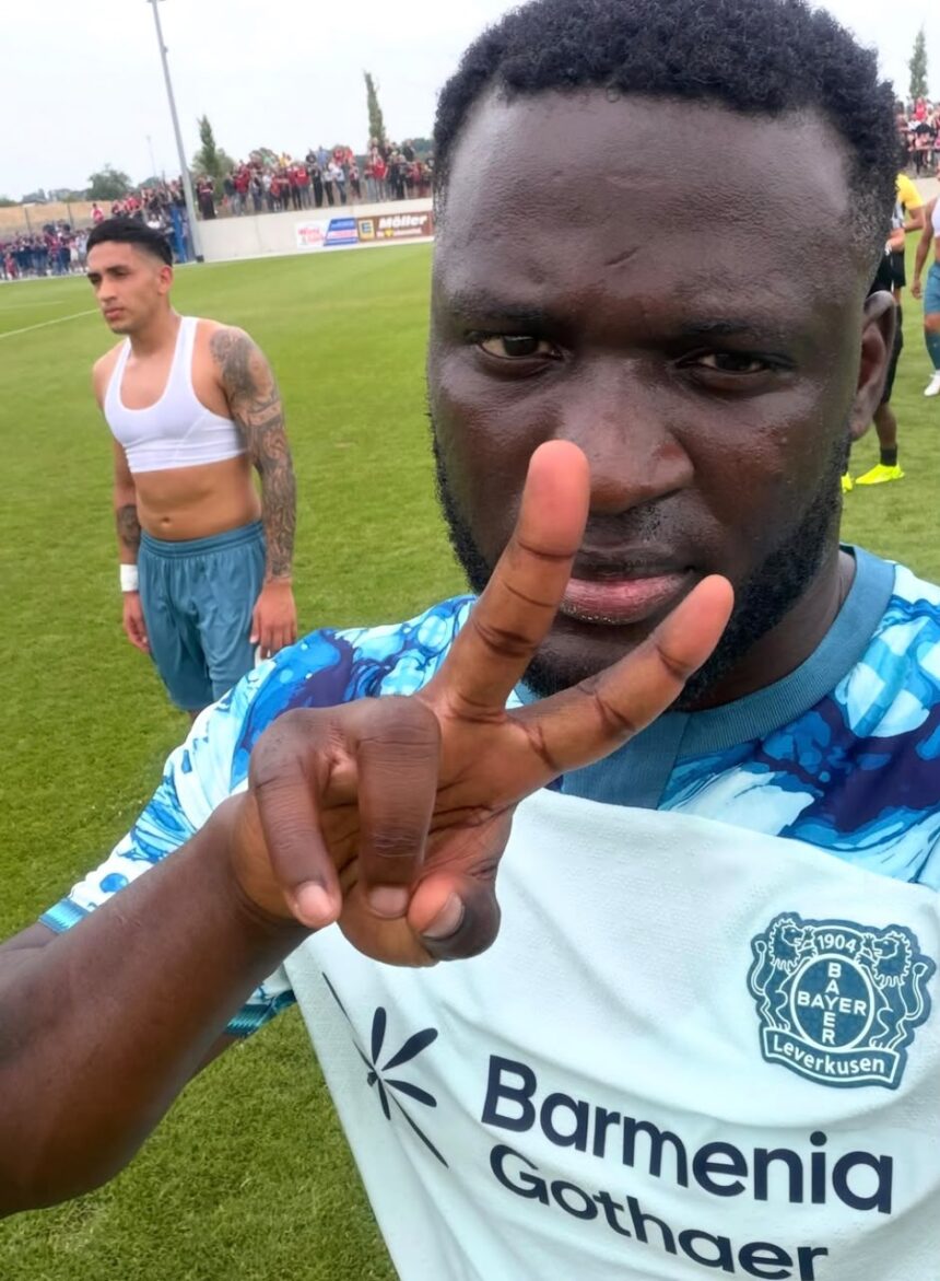 Close up of a footballer on a grassy field wearing a Bayer Leverkusen jersey flashing a peace sign another player in a white top stands behind  trybalnews
