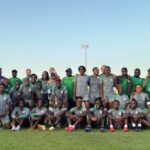 Group photo of a soccer team wearing gray and green uniforms posing on a grass field with coaches standing at the back under a clear sky  trybalnews