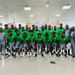 Large group of male athletes in matching green jerseys and gray shorts pose for a team photo in a bright airport terminal with several coaches in gray nearby  trybalnews