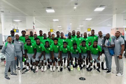 Large group of male athletes in matching green jerseys and gray shorts pose for a team photo in a bright airport terminal, with several coaches in gray nearby.