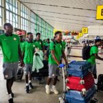 Group of men in green team uniforms walking with rolling suitcases in an airport terminal with bright yellow signage nearby  trybalnews