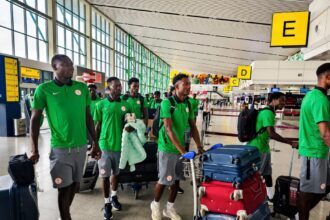 Group of men in green team uniforms walking with rolling suitcases in an airport terminal with bright yellow signage nearby.