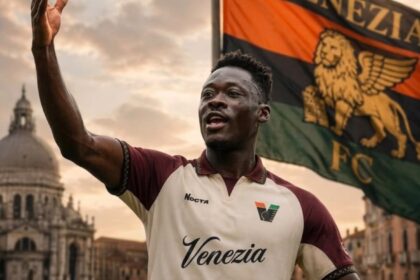 Soccer player in a Venezia FC kit raises an arm in celebration with a flag behind him in a Venetian waterfront setting.