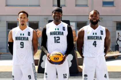 Three Nigerian basketball players posing on an outdoor court in white jerseys (numbers 9, 12, and 4) with the center player holding a basketball.