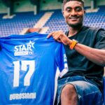 Smiling man in a black T shirt sits in a stadium holding up a blue sports jersey with the number 17 and logos STAR CASINO TV DUROSIMI  trybalnews