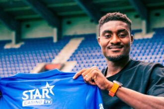 Smiling man in a black T-shirt sits in a stadium, holding up a blue sports jersey with the number 17 and logos (STAR CASINO TV, DUROSIMI).