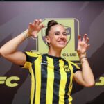 Smiling woman in a yellow and black striped jersey making claw like gesture in front of a club banner logo behind her  trybalnews