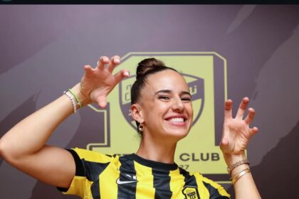 Smiling woman in a yellow and black striped jersey making claw-like gesture in front of a club banner logo behind her.