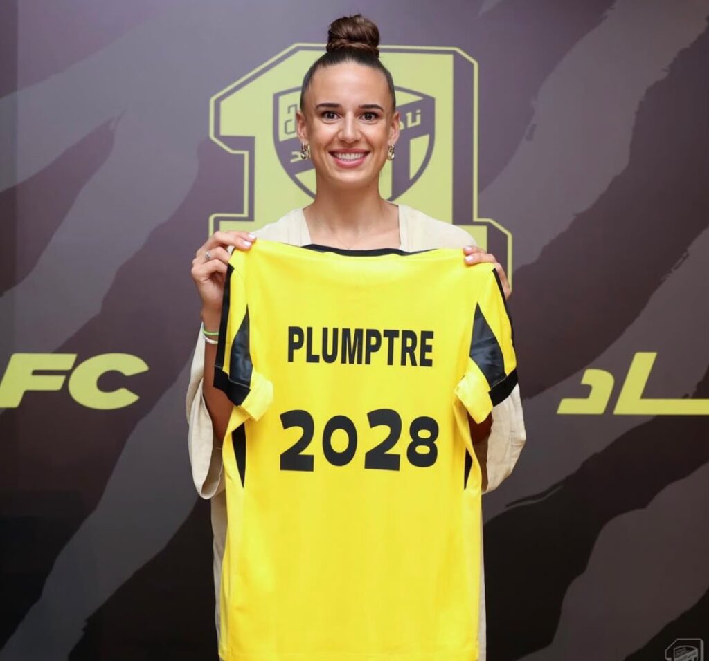 Ashleigh Plumptre Trybal News Smiling woman holds up a bright yellow PLUMPTRE 2028 jersey in front of a sports backdrop trybalnews
