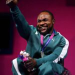 Joyful Paralympic athlete in a teal tracksuit sits in a wheelchair raises a thumb up and shows a silver medal on a purple ribbon around his neck to celebrate a win  trybalnews