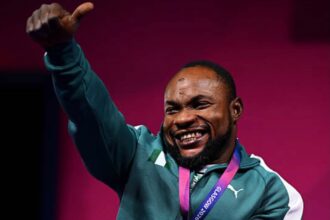 Joyful Paralympic athlete in a teal tracksuit sits in a wheelchair, raises a thumb up, and shows a silver medal on a purple ribbon around his neck to celebrate a win.
