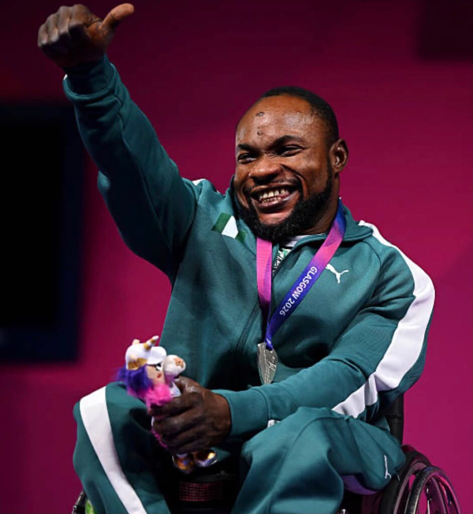 Glasgow Commonwealth Trybal News Joyful Paralympic athlete in a teal tracksuit sits in a wheelchair raises a thumb up and shows a silver medal on a purple ribbon around his neck to celebrate a win trybalnews