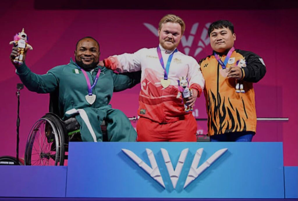Glasgow Commonwealth Trybal News Three medalists on a podium celebrate their victory a wheelchair user on the left a standing athlete in the center and a teammate on the right all wearing medals and holding prizes against a bright pink backdrop with a blue podium featuring a W logo trybalnews