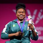 Smiling Black female athlete in a green tracksuit with Nigeria flag wearing a medal and holding a plush unicorn on a pink backdrop  trybalnews