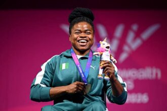 Smiling Black female athlete in a green tracksuit with Nigeria flag, wearing a medal and holding a plush unicorn on a pink backdrop.