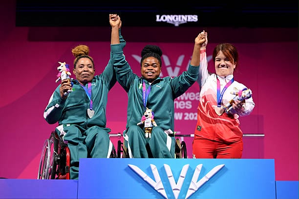 Glasgow Commonwealth Trybal News Three female para athletes on a podium two wheelchair racers on the left with medals one standing on the right all raising their arms in victory trybalnews