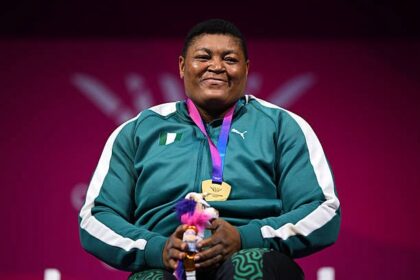 Smiling athlete in a teal tracksuit with a gold medal on a purple ribbon, holding a small stuffed toy against a pink background.