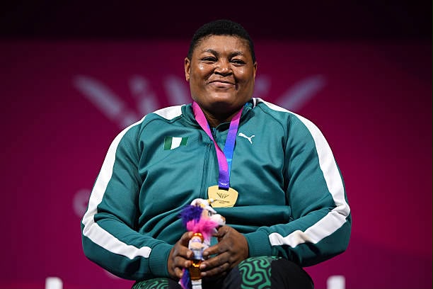 Smiling athlete in a teal tracksuit with a gold medal on a purple ribbon holding a small stuffed toy against a pink background  trybalnews