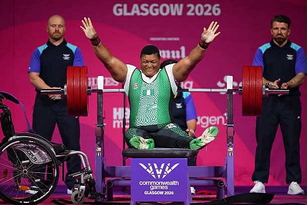 Glasgow Commonwealth Trybal News Paralympic weightlifter on a bench press raising arms in victory bar loaded with red weights two spotters stand behind wheelchair to the left pink backdrop reading Glasgow 2026 trybalnews