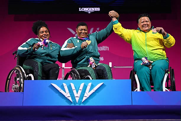 Glasgow Commonwealth Trybal News Three wheelchair athletes celebrate on a podium each holding medals two wear green tracksuits and one wears a yellow green jacket raising their arms in victory on a blue podium with a large W logo trybalnews