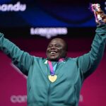 Smiling athlete wearing a teal jacket with a gold medal around the neck raising a trophy in one hand and arms in the air  trybalnews