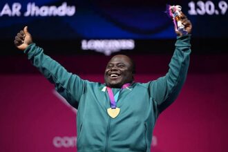 Smiling athlete wearing a teal jacket with a gold medal around the neck, raising a trophy in one hand and arms in the air.