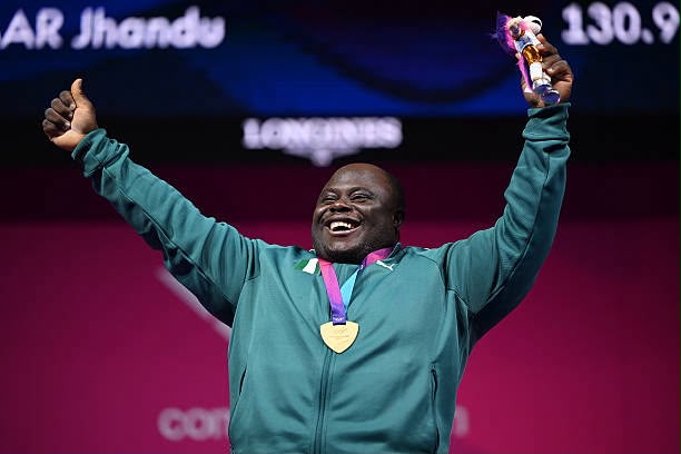 Glasgow Commonwealth Trybal News Smiling athlete wearing a teal jacket with a gold medal around the neck raising a trophy in one hand and arms in the air trybalnews