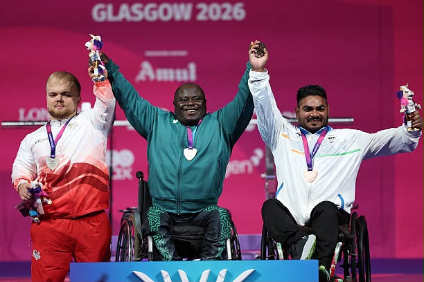 Glasgow Commonwealth Trybal News Three wheelchair athletes on a podium celebrate with medals and trophies at Glasgow 2026 center athlete in teal raises arms high with his teammates nearby trybalnews
