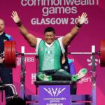 Weightlifter in a green singlet raises arms in victory on a competition platform at the Commonwealth Games Glasgow 2014 pink backdrop behind him  trybalnews