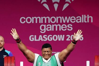 Weightlifter in a green singlet raises arms in victory on a competition platform at the Commonwealth Games Glasgow 2014, pink backdrop behind him.