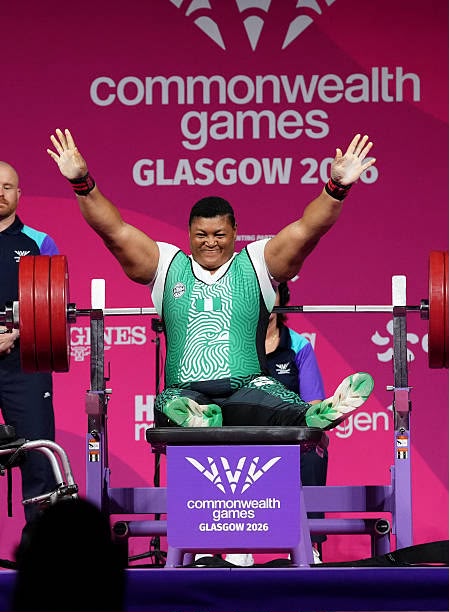 Weightlifter in a green singlet raises arms in victory on a competition platform at the Commonwealth Games Glasgow 2014 pink backdrop behind him  trybalnews