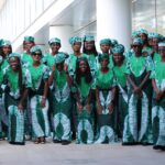Group of people in matching green and white traditional outfits posing outdoors in front of a modern glass building  trybalnews